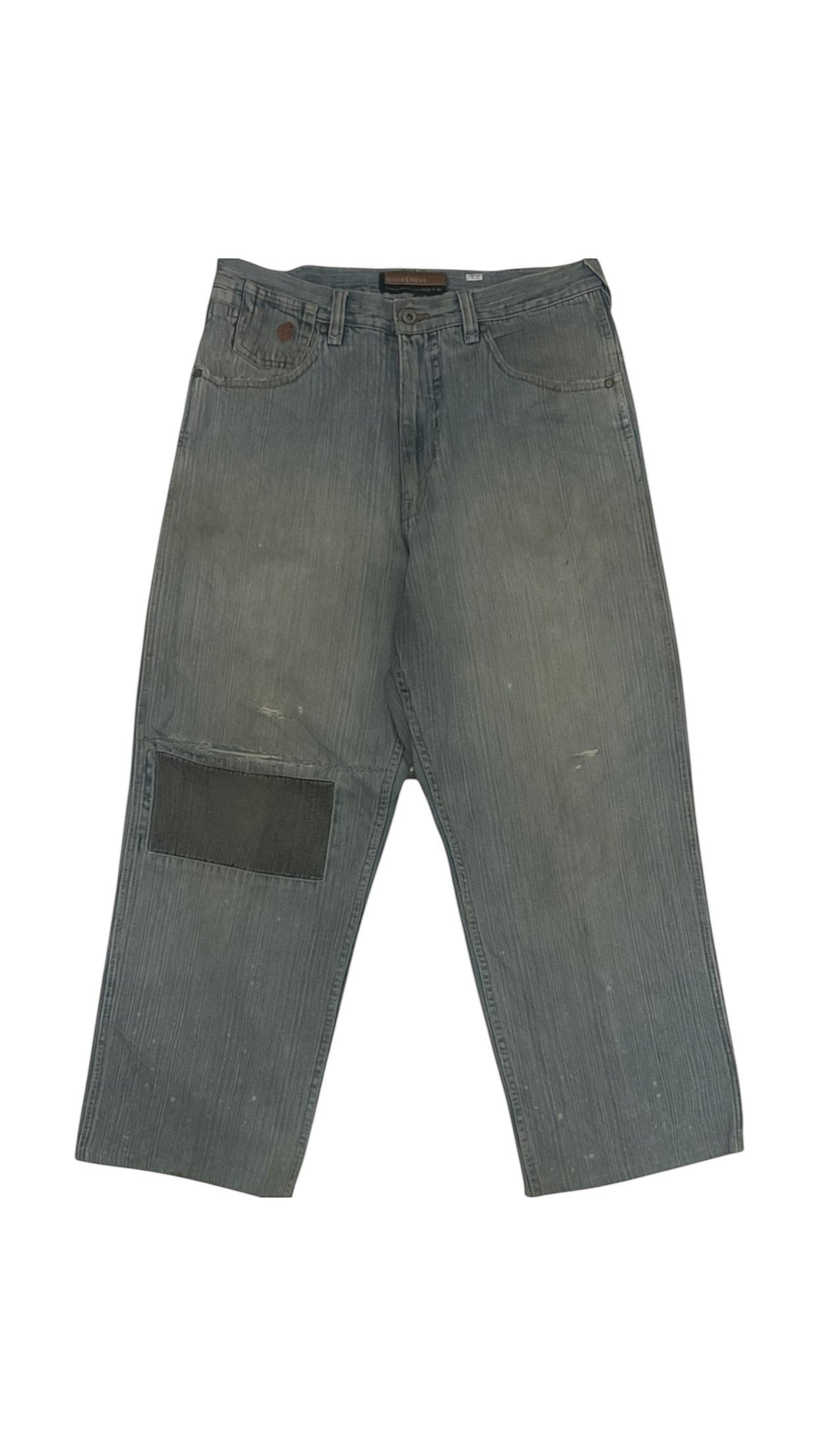 SohoStich Co. distressed faded blue denim jeans with dark grey patchwork on left thigh and paint splatters.